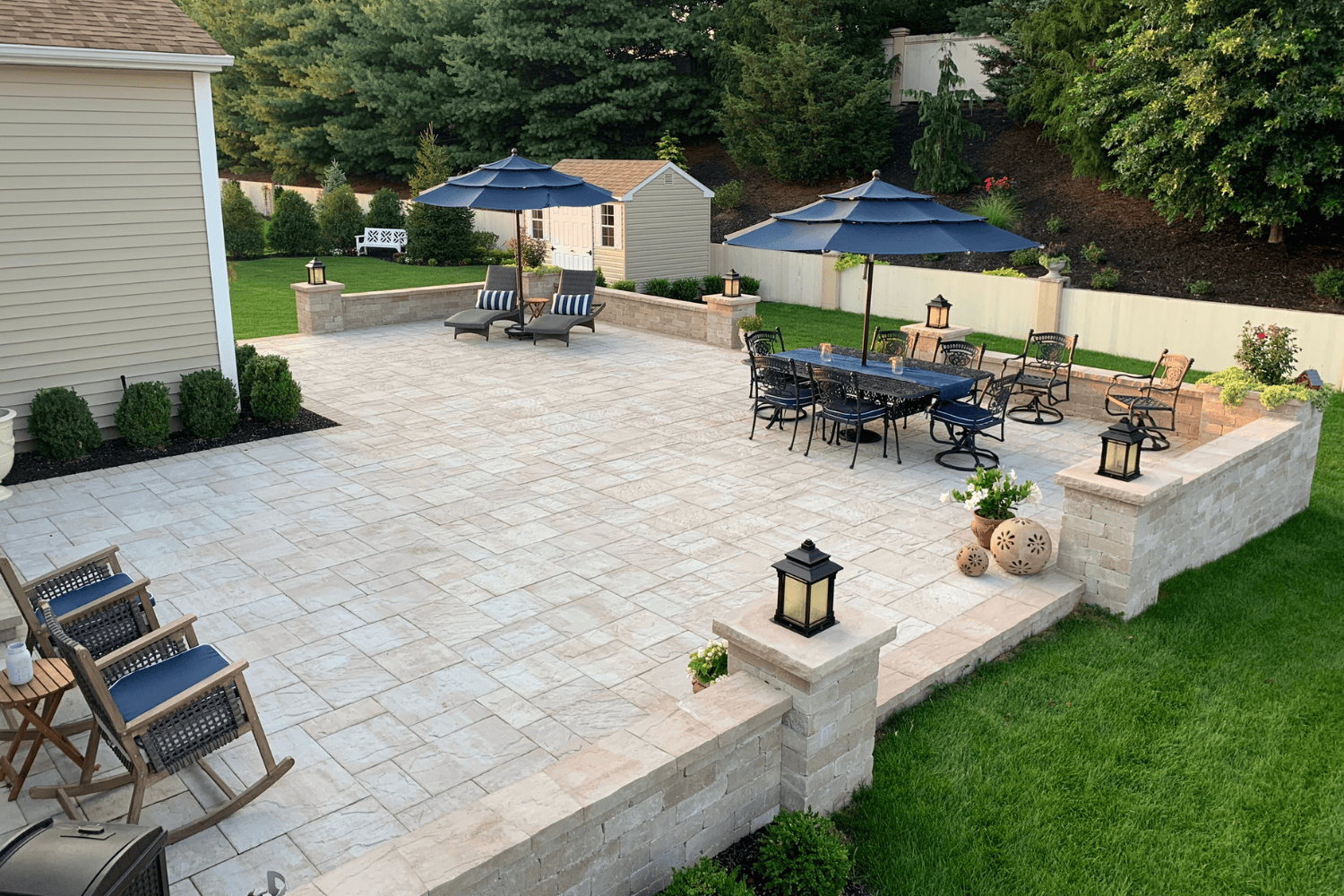 A spacious backyard patio with stone flooring, outdoor seating, dining tables, umbrellas, planters, and lanterns—perfect for relaxing or entertaining. Surrounded by grass and trees, this is one of the most inviting patios you'll find.