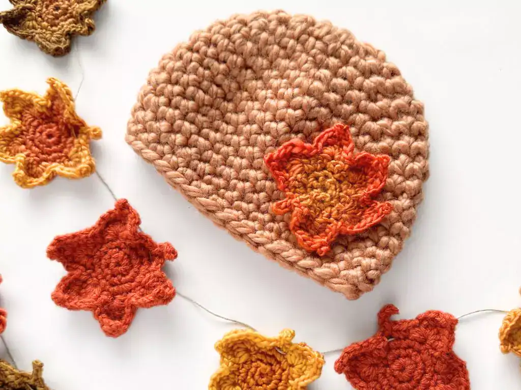 Light brown crocheted beanie with orange maple leaf applique beside a garland of orange and yellow leaves on white background. Free crochet pattern
