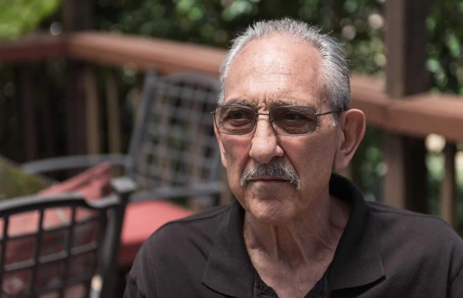 An older man with white hair and glasses, wearing a dark shirt, seated outdoors on a porch with trees behind him.