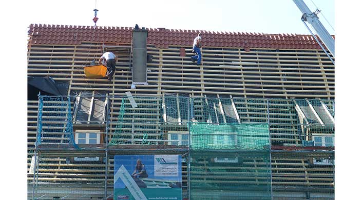 Ein Bauarbeiter arbeitet ohne sichtbare Sicherung auf einem Hausdach, das sich im Bau befindet und teilweise abgedeckt ist. Baugerüste und Netze sind zu sehen.