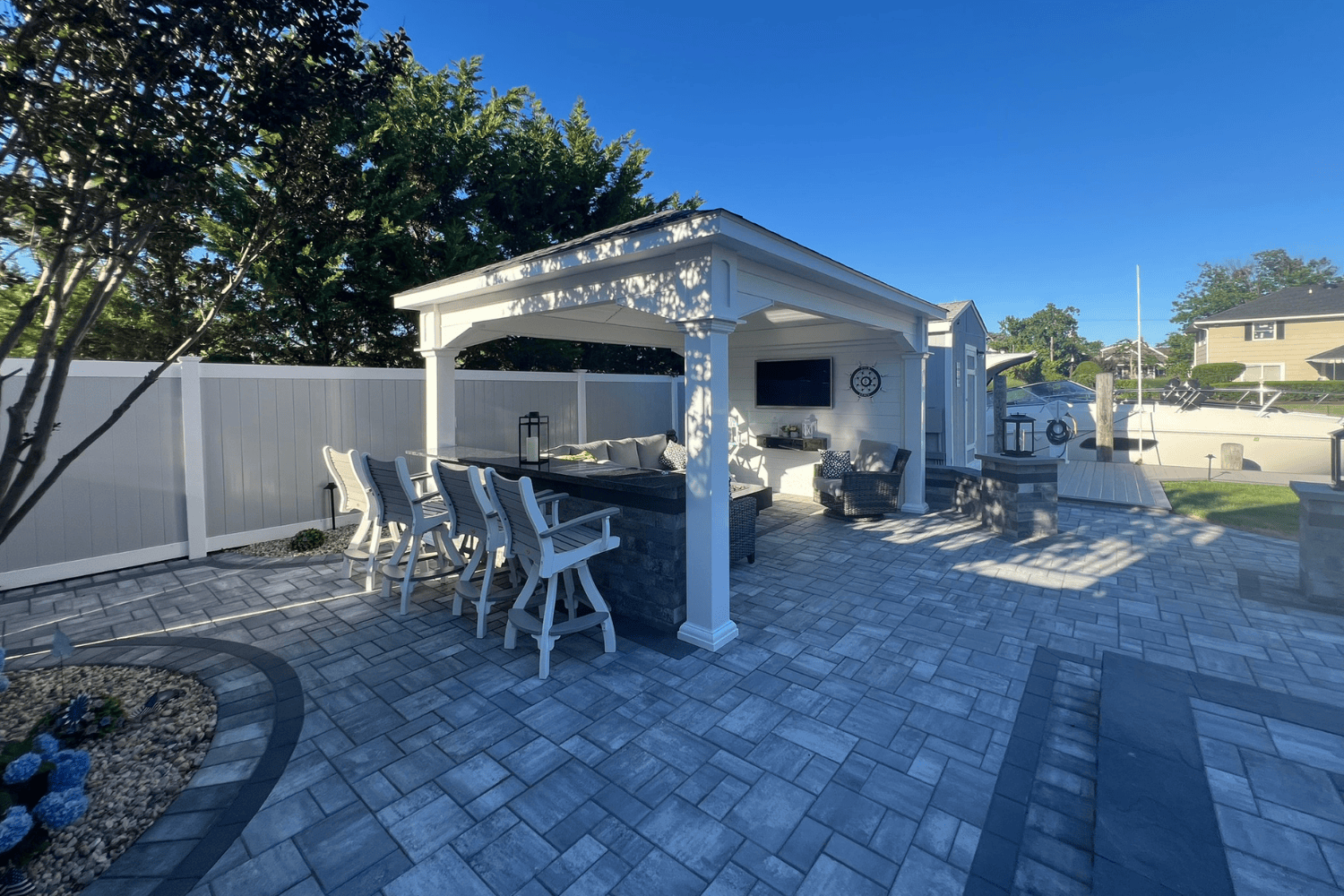 A backyard patio features a covered seating area with high chairs and a TV beneath stylish pergolas, paved flooring, and a white fence, all under a clear blue sky.