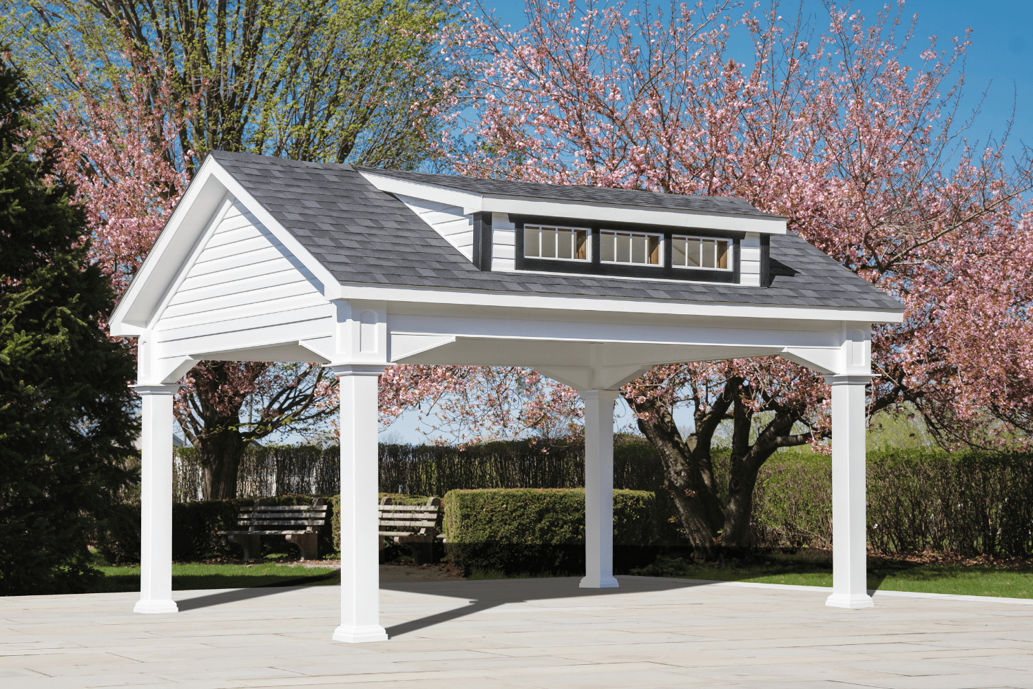 A white, four-posted pavilion with a gray shingled roof stands on a stone patio, surrounded by blooming trees and trimmed hedges under a blue sky—an inviting space among elegant pavilions and pergolas.
