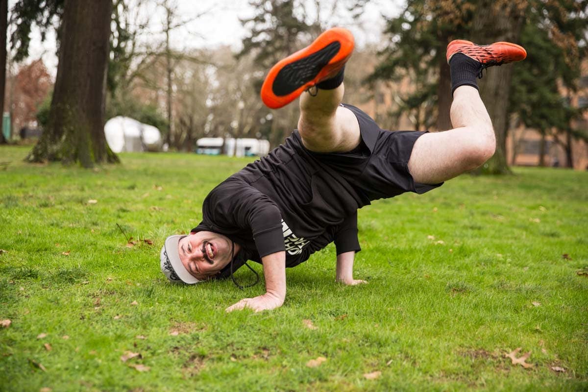 A man wearing athletic clothes and orange shoes balances on one hand and his head in a grassy park, with one leg raised and bent. Trees are visible in the background.