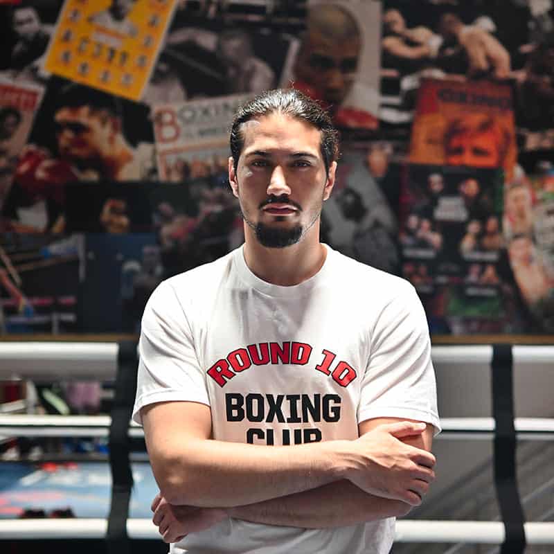 A formidable presence at Round 10 Boxing Club: A determined man, his long hair pulled back and beard well-kept, commands the room with arms crossed in resolute confidence. Clad in the signature Round 10 t-shirt, he stands amid the iconic walls of our Dubai gym—where legendary boxing posters and a relentless spirit inspire every fighter who enters.