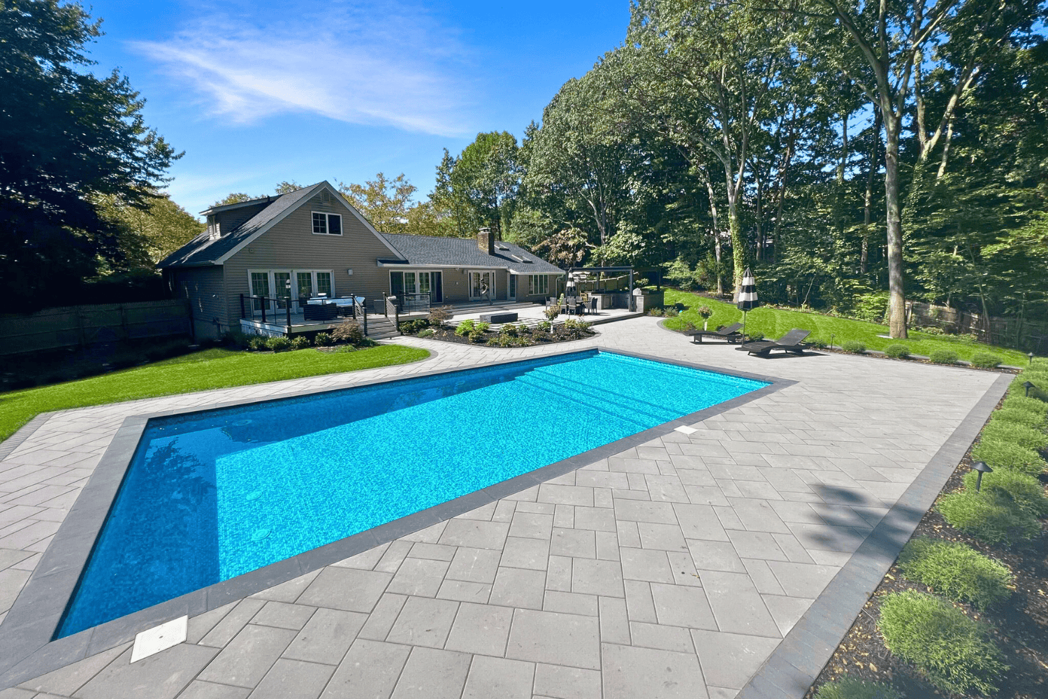 Rectangular outdoor swimming pool surrounded by stone patios, lounge chairs, and landscaped greenery, with a house and trees in the background under a clear sky.