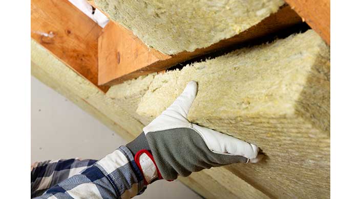 Eine Person mit Arbeitshandschuhen installiert eine gelbe Dämmplatte zwischen Holzbalken an einer Wand.