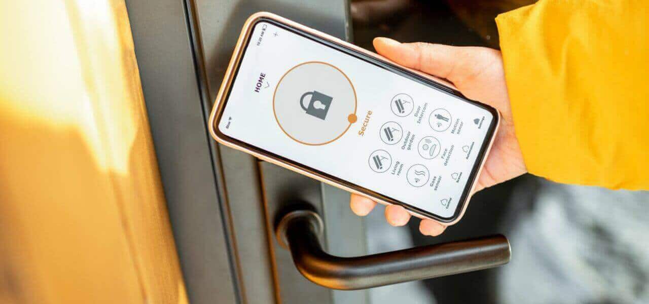 A person wearing a yellow jacket is using a smartphone to unlock a door with a smart lock application. The phone screen displays the app’s interface with a large lock icon and several options below it, making them look like a digital locksmith. The door has a black, modern handle.