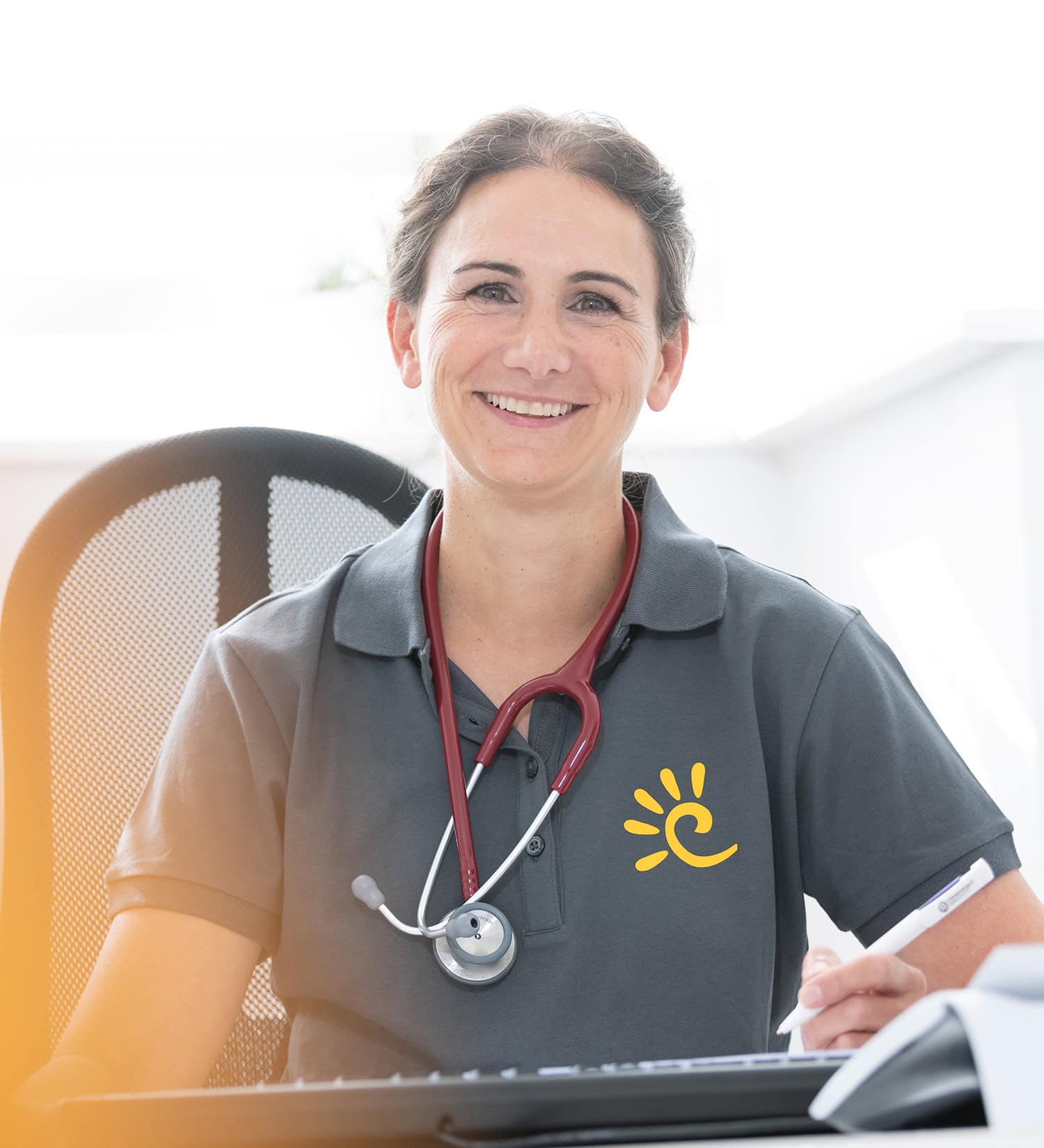 Lächelnde Ärztin sitzt auf einem Bürostuhl und hat ein rotes Stethoskop um den Hals.
