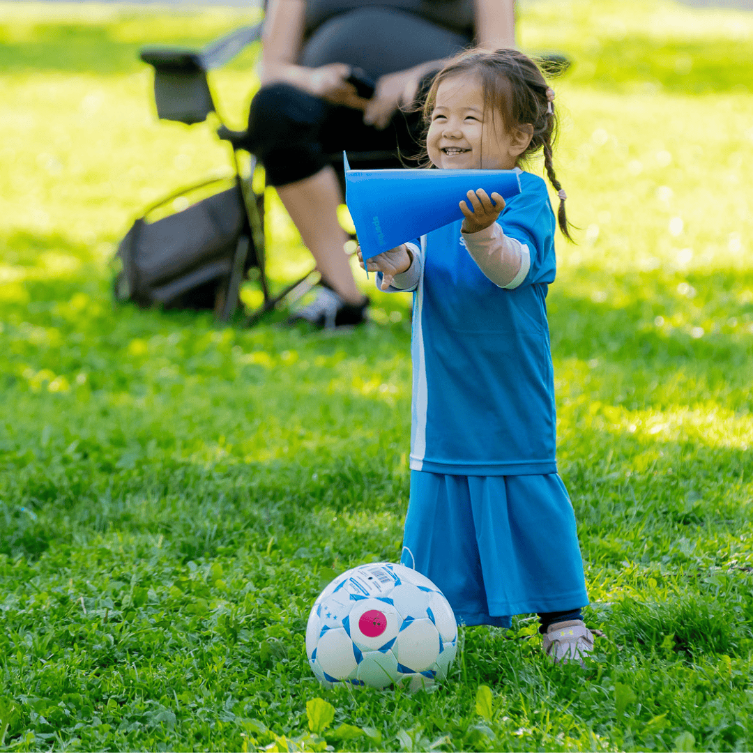 Young child in a blue Sportball uniform smiles while holding a blue cone beside a soccer ball during an outdoor class.