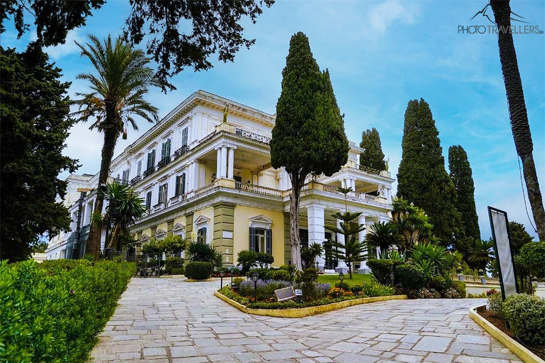 Bäume, Palmen und Steinterrassen umgeben die in weiß- und gelbtönen gestrichene Residenz Achilleion