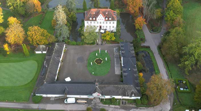 Luftaufnahme eines weißen Herrenhauses mit rotem Dach, vorne ein runder Garten mit Statuen, umgeben von Bäumen und Wegen.