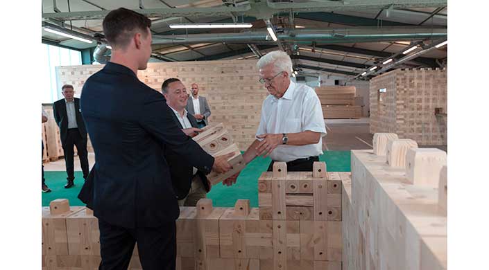 Ein älterer Mann hilft, große Holzbausteine zu stapeln, während ein weiterer Mann zusieht. Die Szene findet in einem Innenraum mit grünem Boden statt.