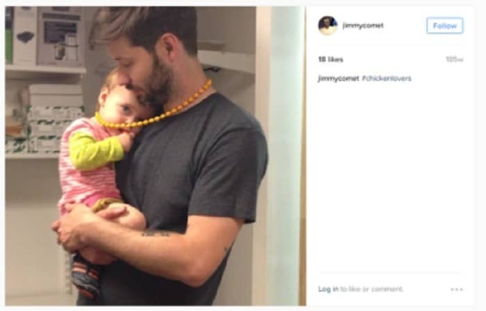 A father holds a baby, both facing the camera. The baby is wearing a pink and green outfit and is playing with a yellow necklace. The man is wearing a dark gray t-shirt. This image may be related to child abuse scandals and elite networks.