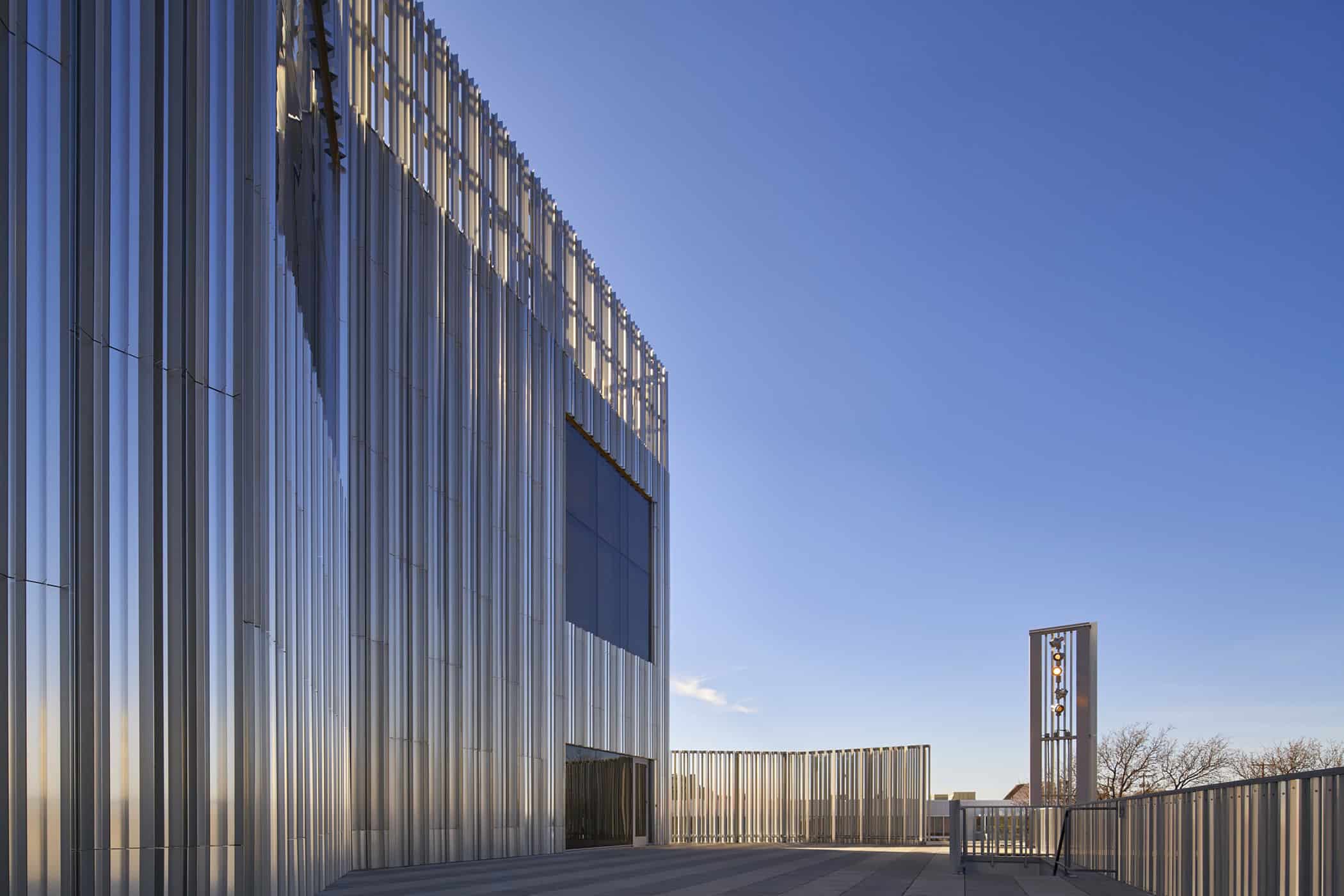 Close-up exterior view of the Oklahoma Contemporary Arts Center, showcasing the vertical aluminum fins and reflective façade design under a clear blue sky.