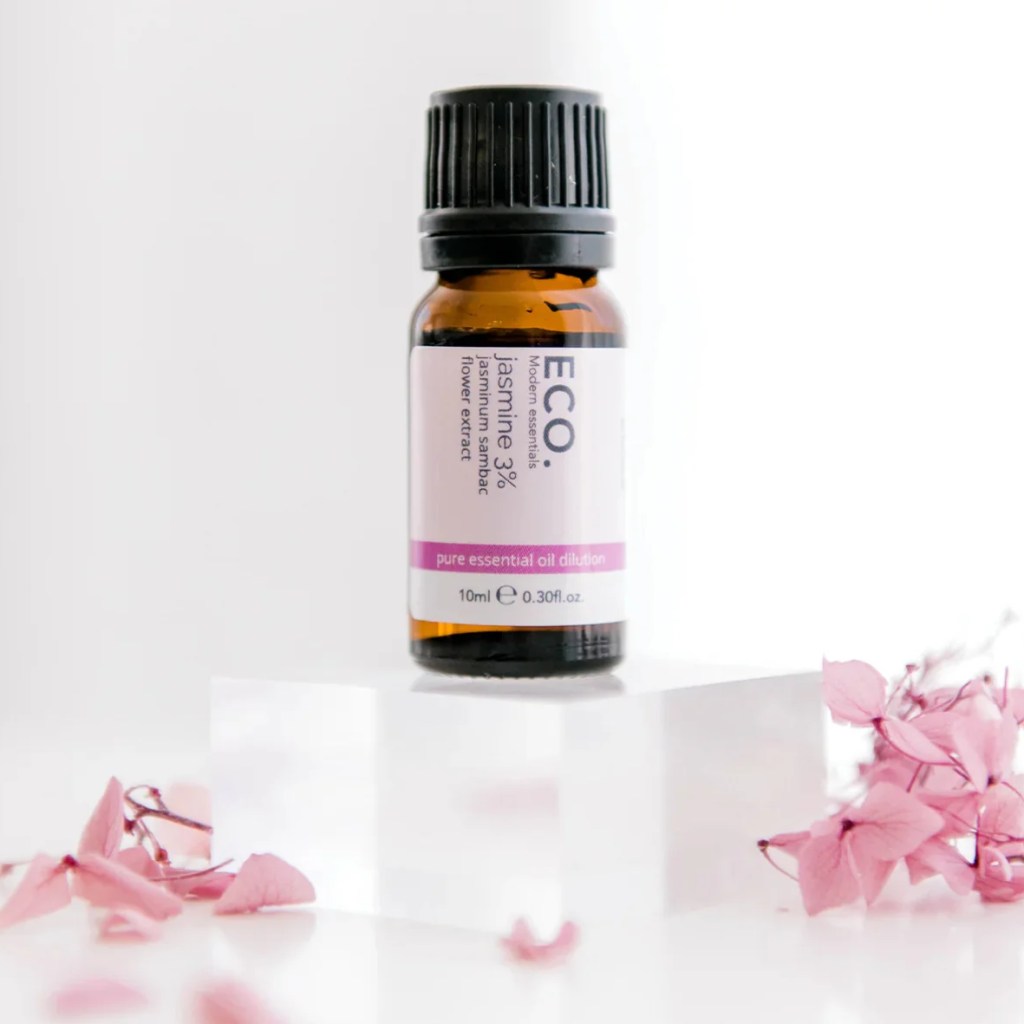 A small brown bottle of Jasmine Essential Oil rests on a clear acrylic block, surrounded by pink dried flower petals against a white background.