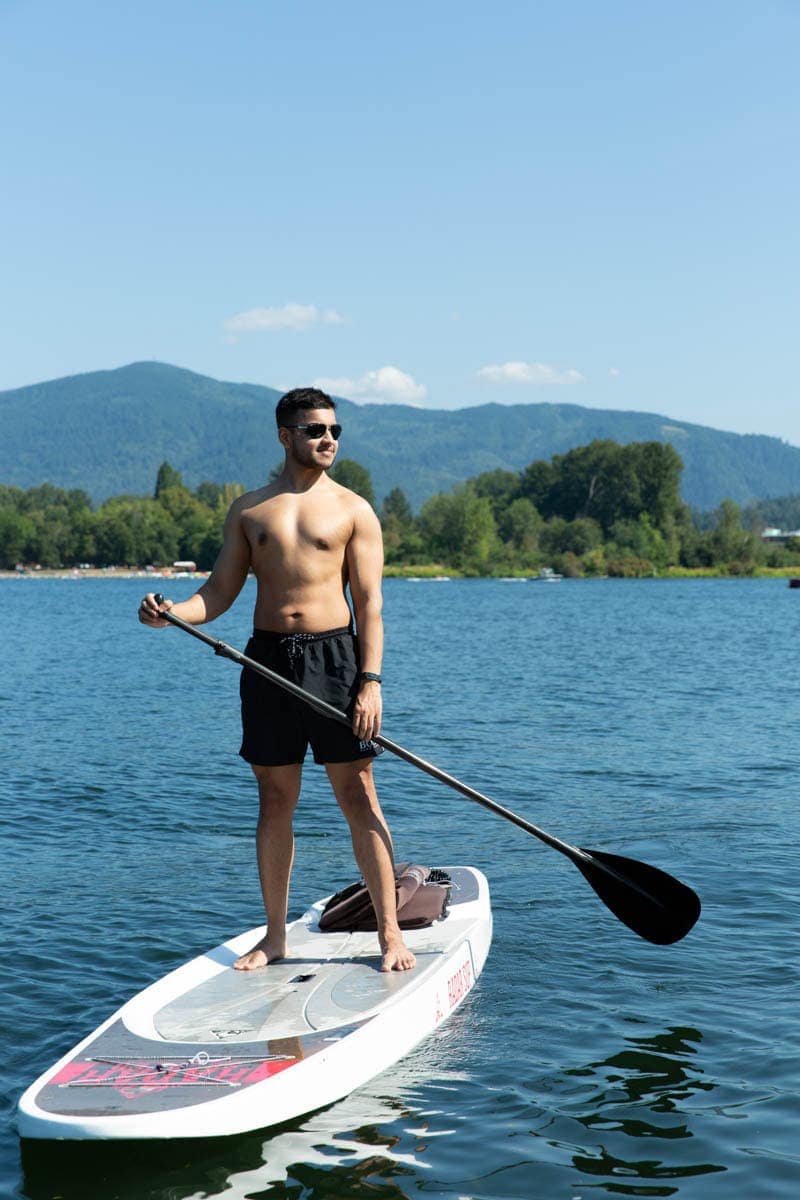 Man paddleboarding on a calm lake, wearing sunglasses and shorts, with green hills and clear blue sky in the background.