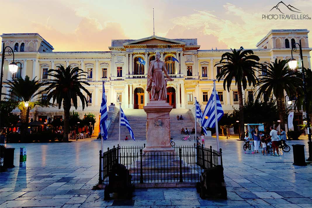 Vor dem weißen, neoklassizistischen Verwaltungsgebäude am Miaouli-Platz prangt ein steinernes Denkmal umrahmt von griechischen Flaggen
