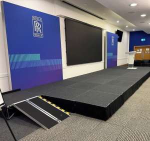 wheelchair-ramp-for-staging-access - NexGen Portable Staging & Dancefloors Conference stage with blue Rolls-Royce banners, a large black screen, a podium, and a portable ramp at the front.