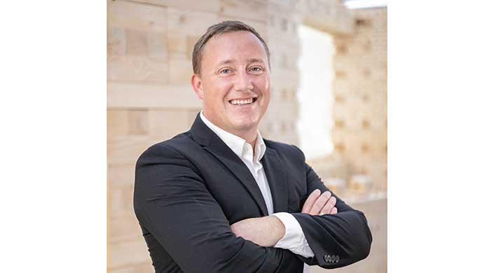 Ein lächelnder Mann im Anzug steht mit verschränkten Armen vor einem hellen, unscharfen Hintergrund.