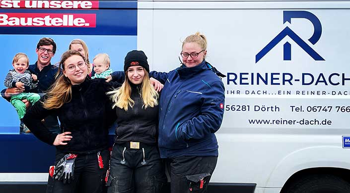 Zwei Frauen in Arbeitskleidung stehen nebeneinander, eine trägt eine Brille und beide lächeln, während sie vor einer weißen Wand posieren.