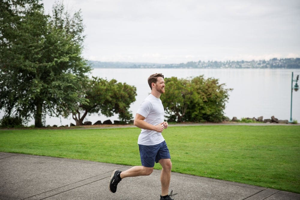 A man in a white shirt and blue shorts jogs on a paved path near a grassy area by a lake, with trees and water in the background.