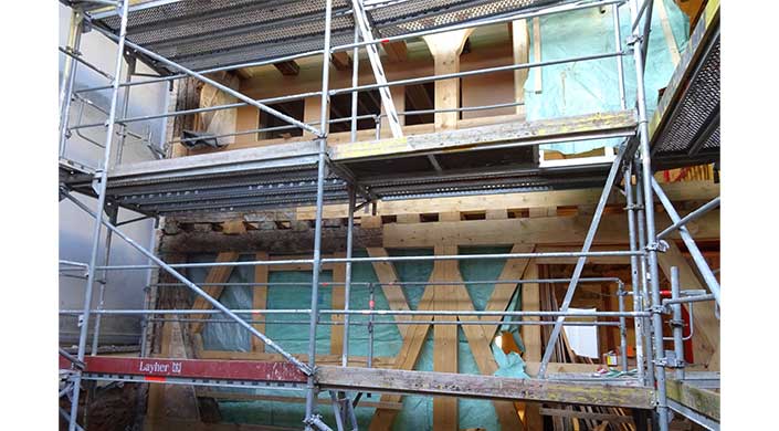 Gerüst und sichtbare Holzbalken einer Fachwerkhaus-Baustelle, teilweise abgedeckt mit grüner Folie, zeigen laufende Bauarbeiten und traditionelle Bauweise.