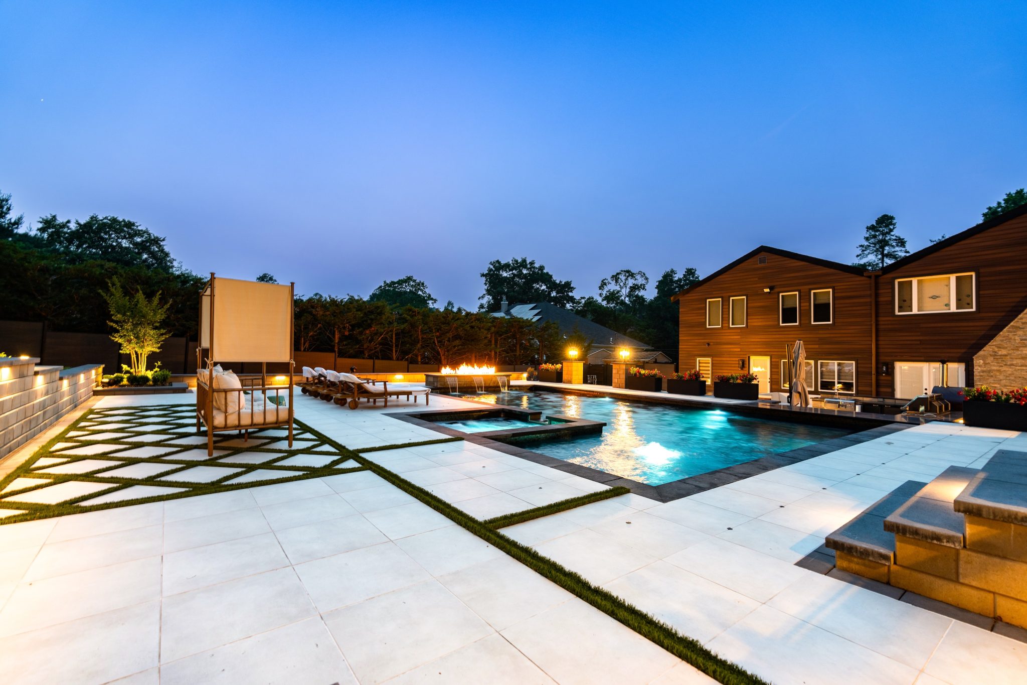 Modern backyard with a rectangular swimming pool, lounge chairs, fire feature, and landscaped patio area beside a two-story house at dusk.