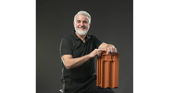 Älterer Mann mit grauem Haar und Bart lächelt, trägt ein schwarzes Poloshirt und hält ein orangefarbenes Objekt vor dunklem Hintergrund.