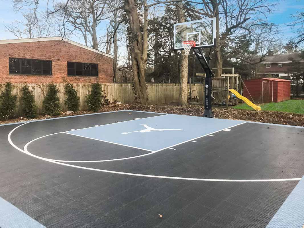 Outdoor sports court with a black and blue color scheme, featuring a hoop and backboard. The court is situated in a backyard in Huntington, surrounded by trees with a slide to the right for extra fun.