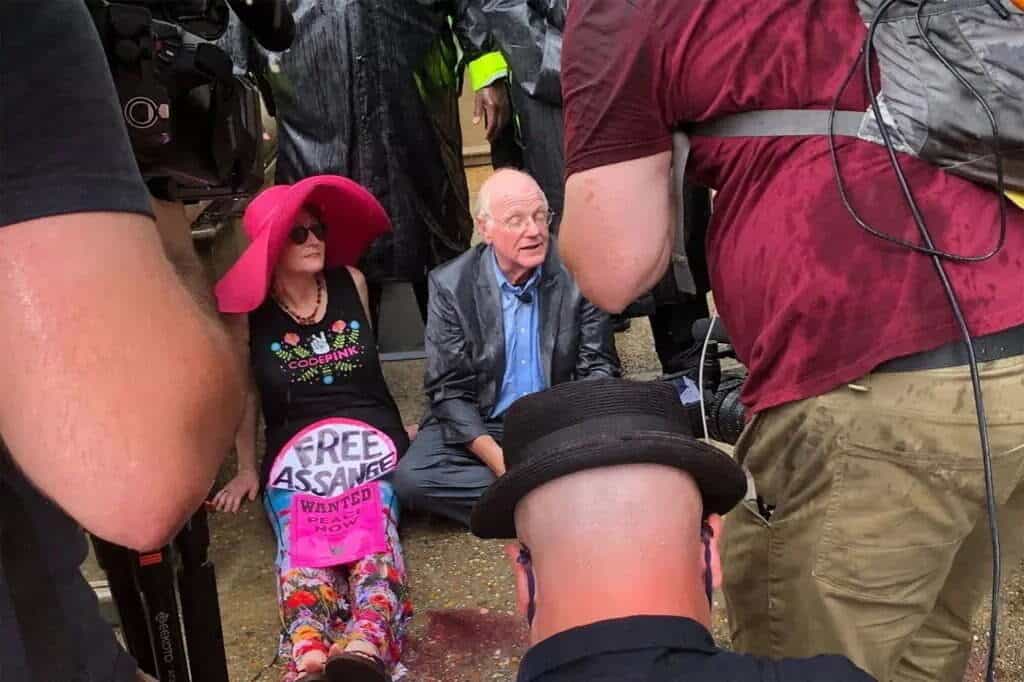 Ben Cohen, co-founder of Ben & Jerry's, sits during a rainy Julian Assange protest in DC. A CODEPINK activist holds a "Free Assange" sign. Other protesters and media surround them, blocking the DOJ entrance. The protest highlights the fight for press freedom.