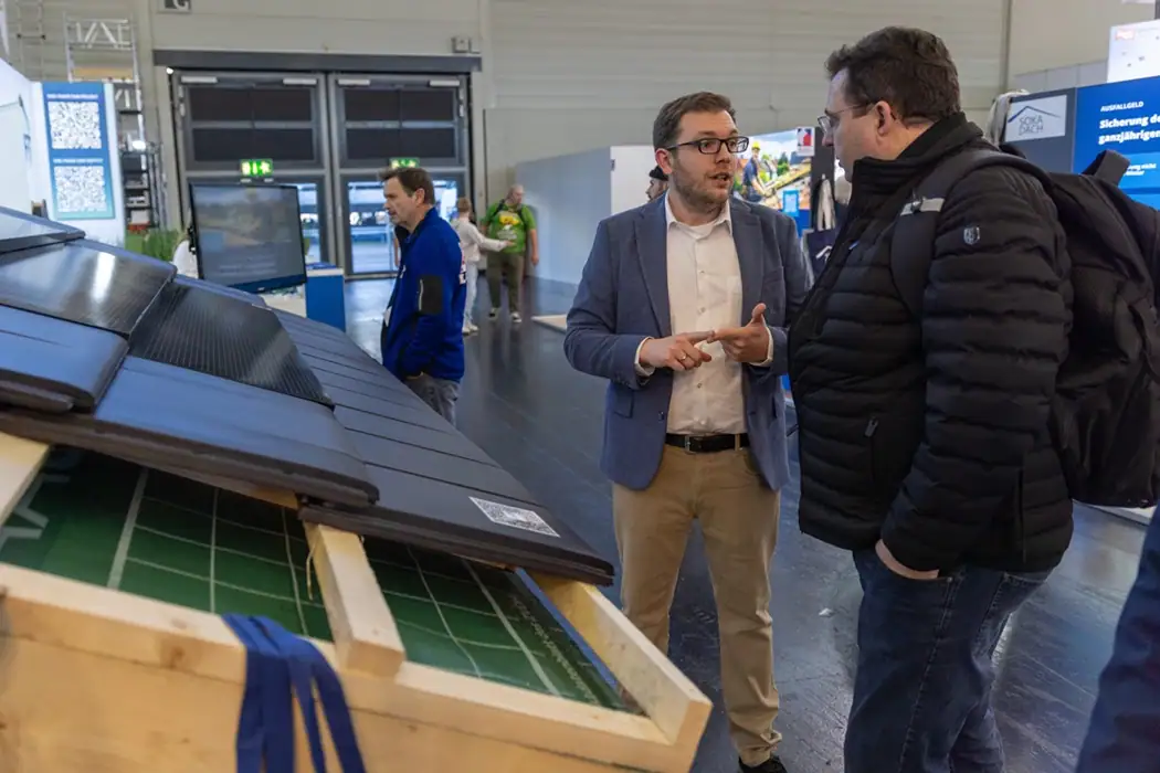 Jan Redecker (links) diskutieren mit einem Besucher er auf der Messe Dach+Holz in Köln neben einer Solarziegel-Dachinstallation, während andere Besucher und Stände im Hintergrund zu sehen sind.