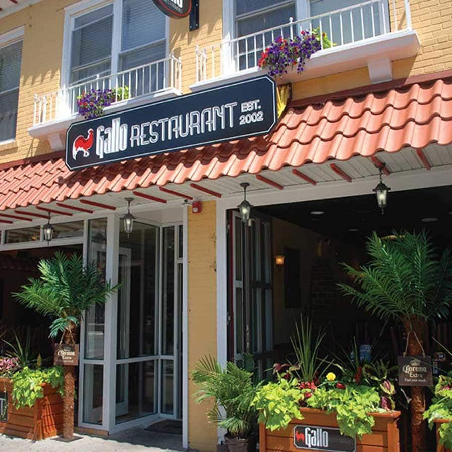 Exterior view of Gallo Restaurant with red tile roof, potted plants, and outdoor seating under a covered patio. Signs display the restaurant's name and “Est. 2002.”.