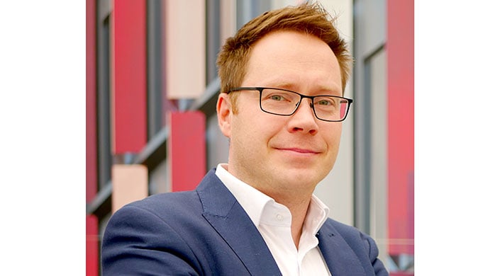 Ein Mann mit Brille und braunen Haaren trägt einen blauen Anzug und weißes Hemd, steht vor einem modernen Gebäude, nur die rechte Gesichtshälfte ist sichtbar.
