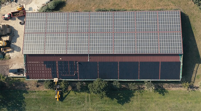 Solarmodule auf dem Dach eines Gebäudes, umgeben von Gras und Bäumen, aus der Vogelperspektive fotografiert.