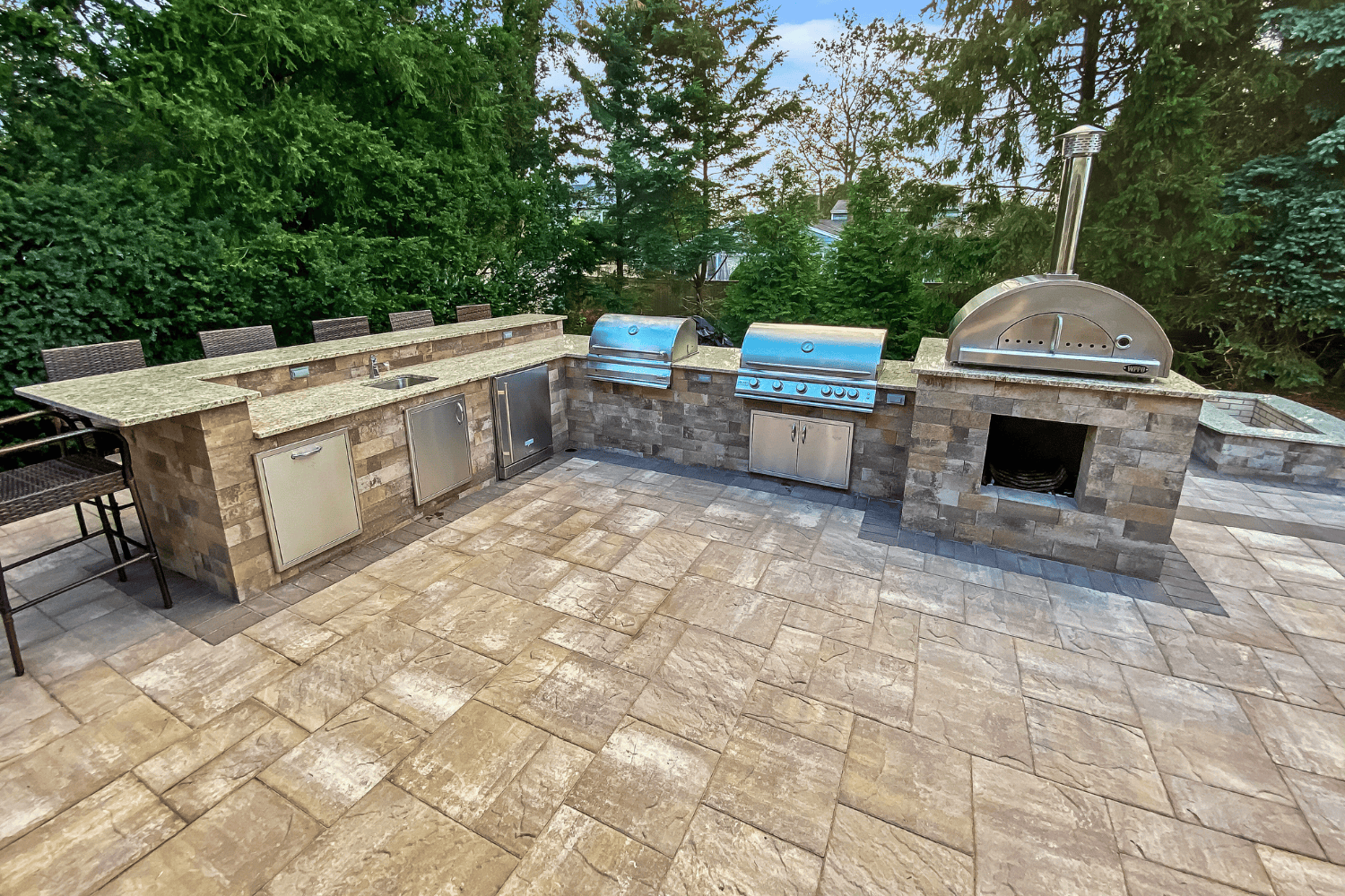Outdoor kitchen with stone countertops, stainless steel appliances, a grill, pizza oven, sink, and bar seating—this stunning example of Outdoor Kitchens sits on a tiled patio framed by lush trees.