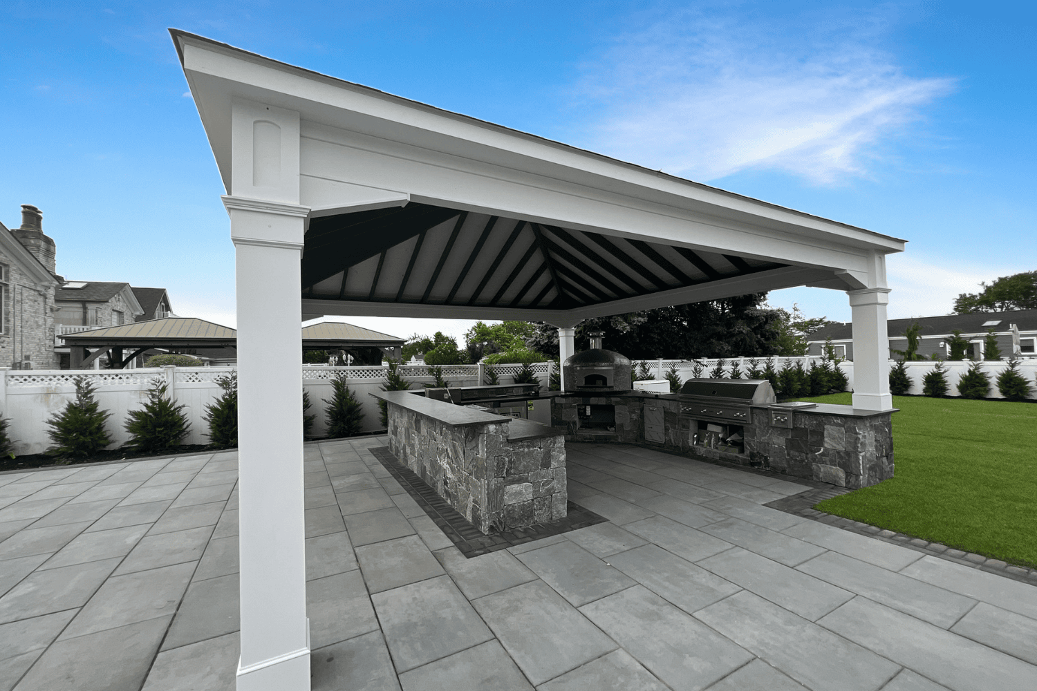 Covered outdoor kitchen with stone countertops, built-in grill, pizza oven, and stainless steel appliances on a stone patio, all sheltered under elegant pavilions and surrounded by lush lawn and landscaping.