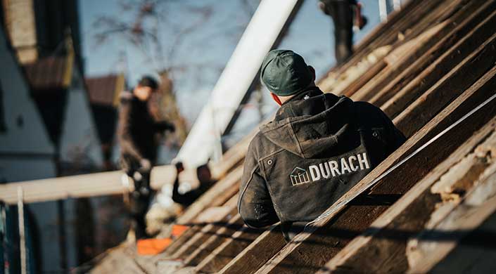 Ein Arbeiter mit grüner Mütze und dunkler Jacke arbeitet auf einem Dach aus Holzlatten, im Freien bei Tageslicht.