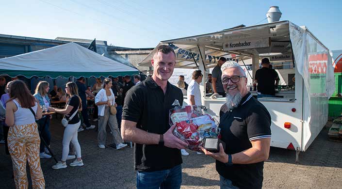 Ein Mann mit kurzem Haar hält einen Geschenkkorb im Freien, umgeben von weiteren Menschen bei sonnigem Wetter.