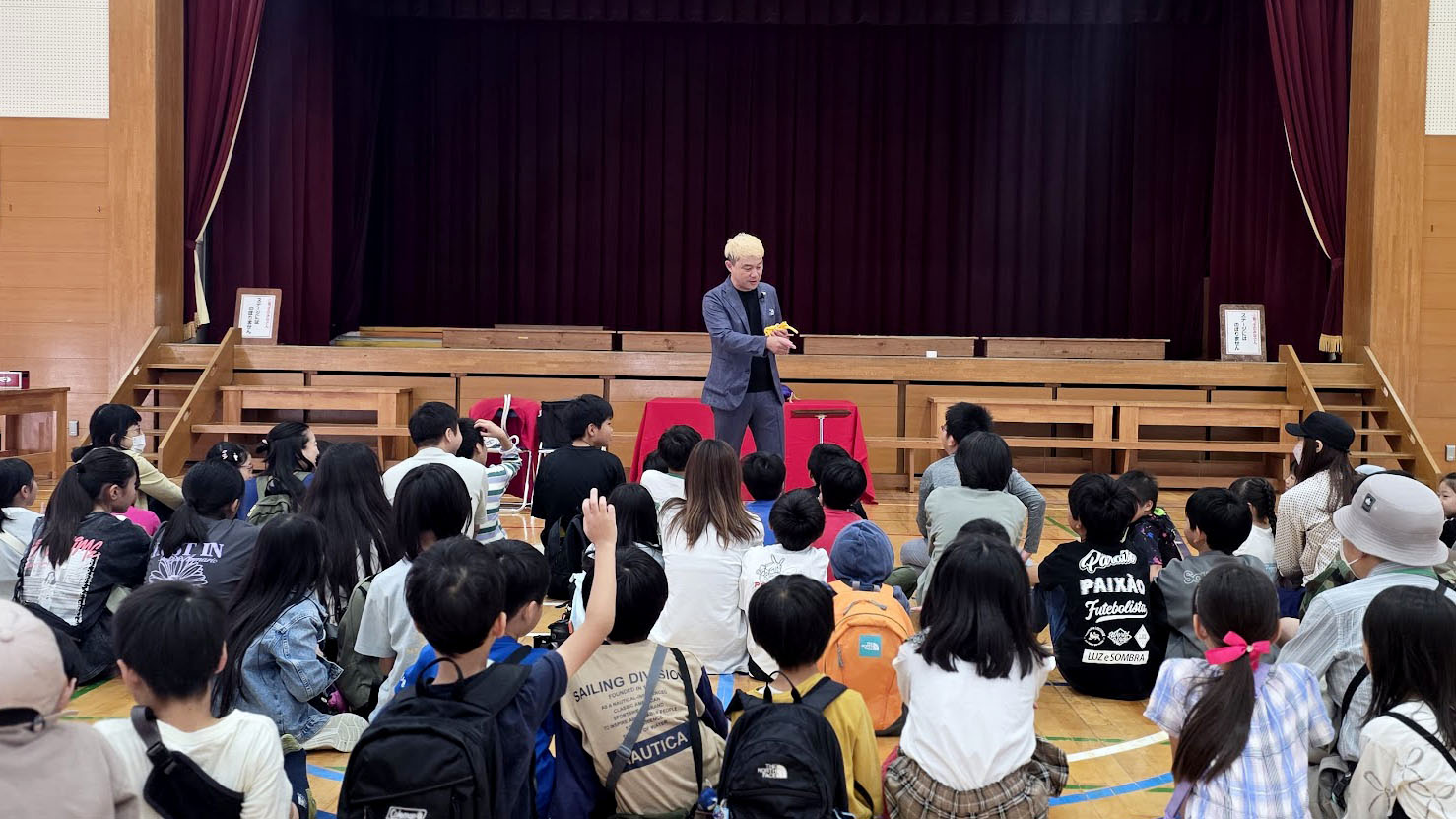 金沢市小坂小学校で2年連続マジックショー!昨年との違いに感じた「文化を育てる」ということ 1 体育館のステージ前でマジックを披露するマジシャン・コンプレッサー。床に座ったたくさんの子供たちと保護者がショーに注目している。