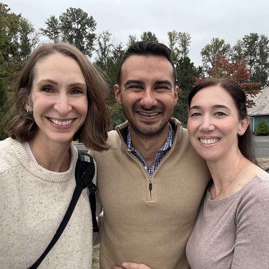 Three people stand outdoors, close together and smiling at the camera, with trees and a house visible in the background.