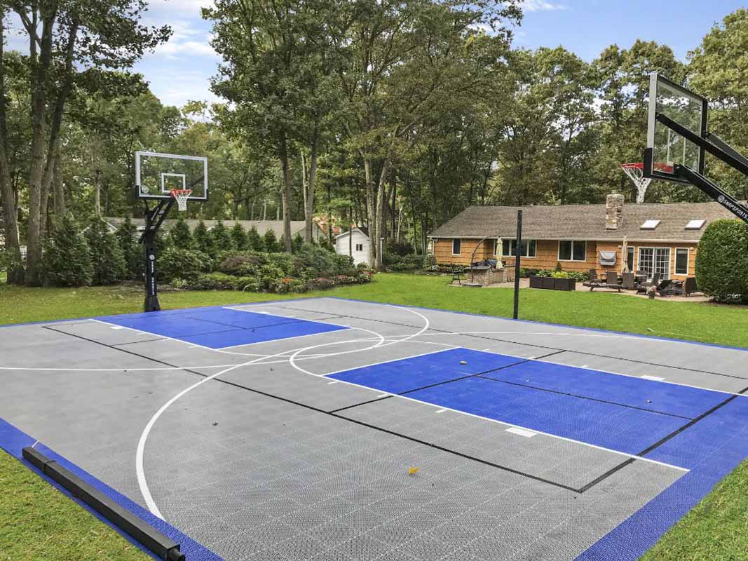 Outdoor basketball court with blue and gray tiles, surrounded by trees. In the background, there is a house with a patio and lawn furniture—an ideal sports court backyard installation in Huntington.