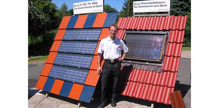 Ein Mann steht vor einer solarbetriebenen Anlage mit Solarpanelen und Dachziegeln, trägt weißes Hemd und schwarze Hose, draußen bei sonnigem Wetter.