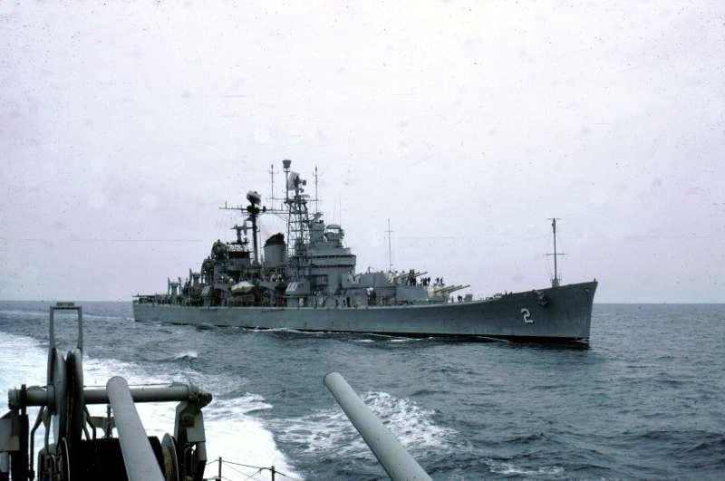 A gray US Navy cruiser underway at sea, seen from another ship, with its forward gun turrets and tall masts against a hazy sky.