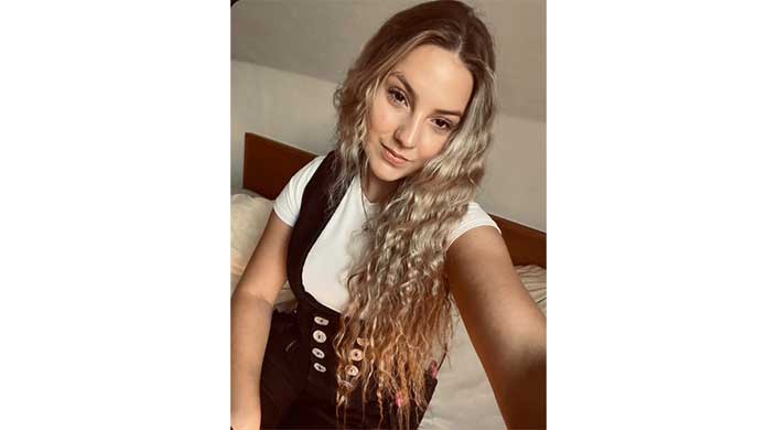 Junge Frau mit langen, lockigen, blonden Haaren trägt weißes Shirt und schwarze Weste, blickt lächelnd in die Kamera.