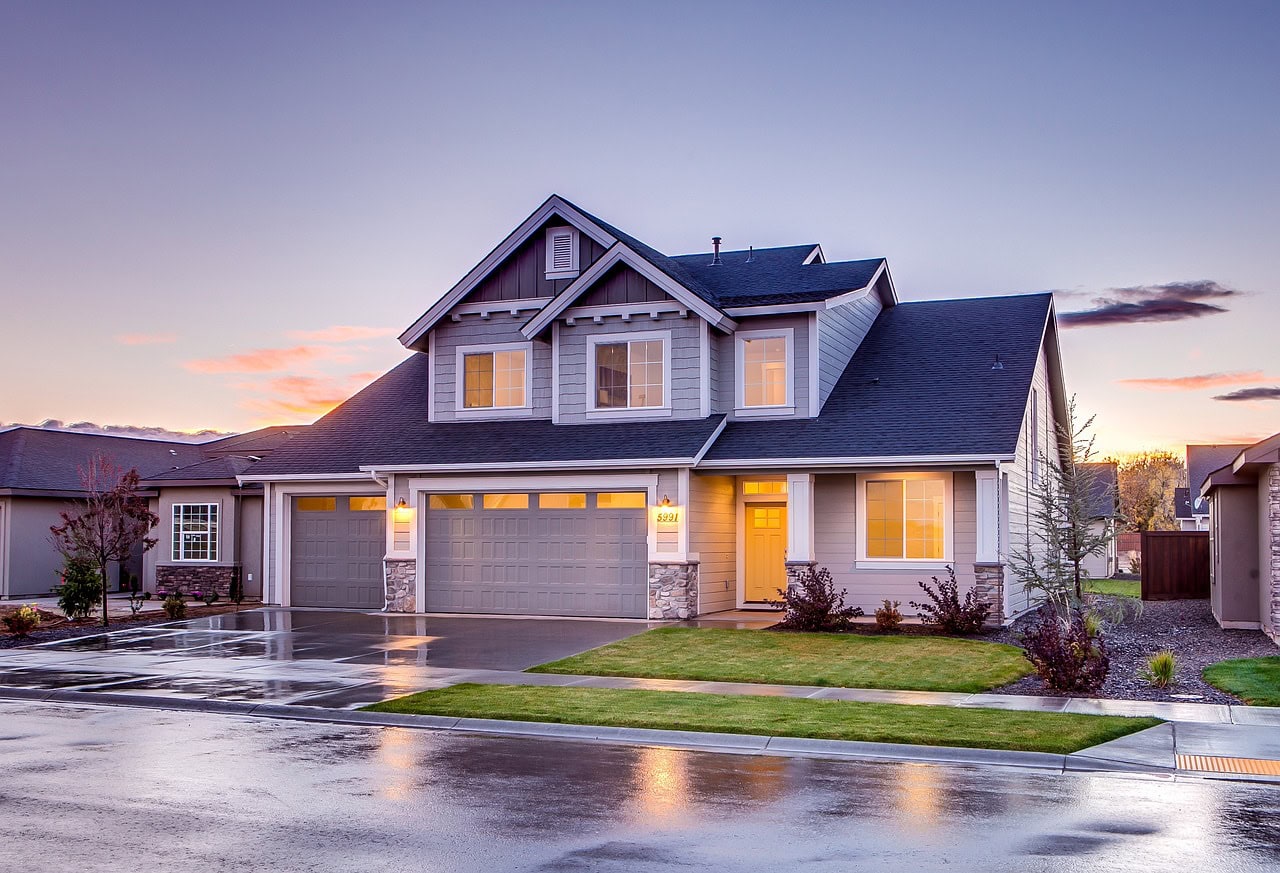 Suburban two-story house with a double garage and landscaped yard at dusk, wet driveway reflecting the pink sky.