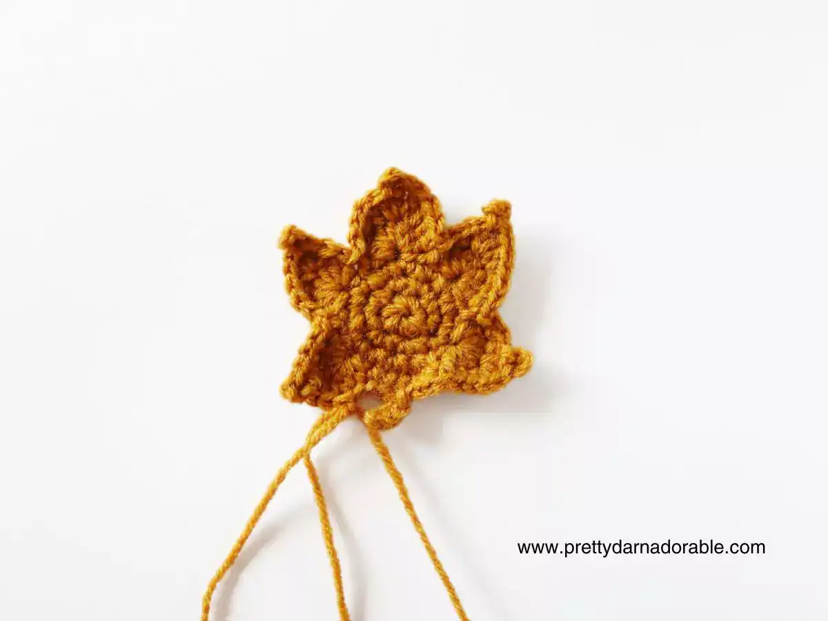 Mustard yellow crochet maple leaf with two yarn tails on white background; www.prettydarnadorable.com in bottom right corner. Free crochet pattern