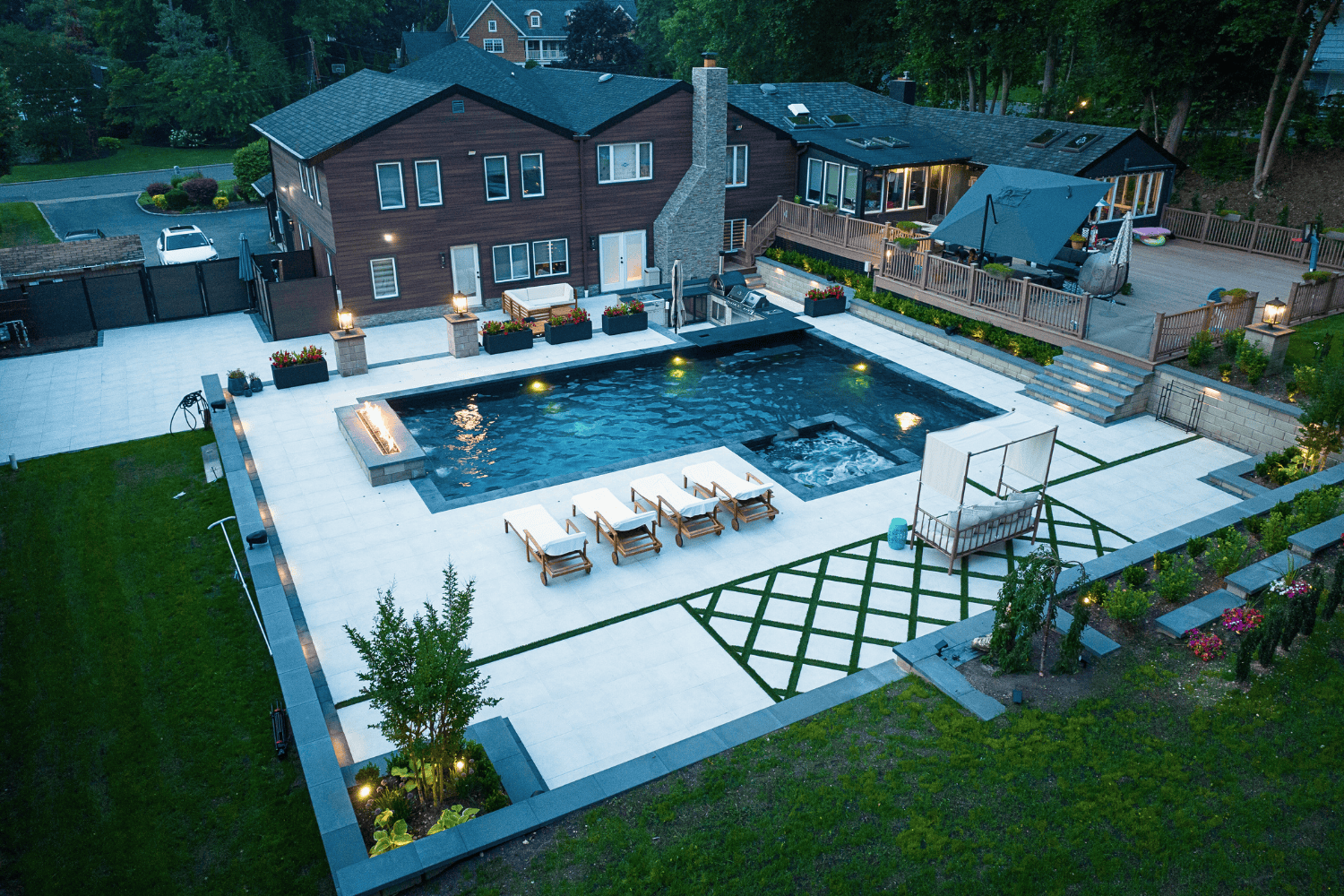 Aerial view of a modern backyard featuring a large rectangular pool, patios with lounge chairs, hot tub, patio seating, and landscaped greenery beside a two-story house.
