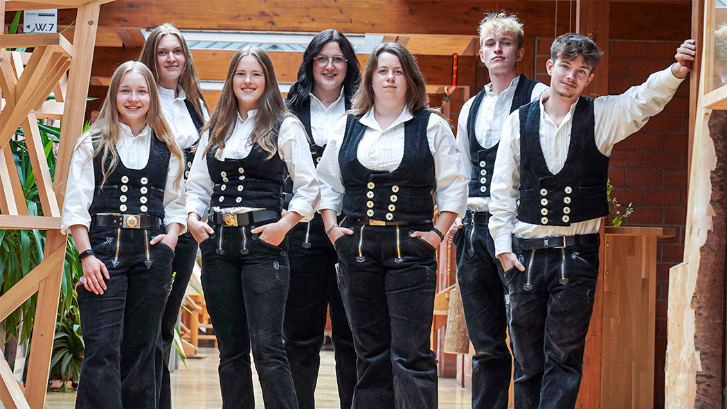 Zwei Frauen in traditioneller Handwerkskleidung mit weißen Hemden und schwarzen Westen stehen nebeneinander in einem Innenraum.
