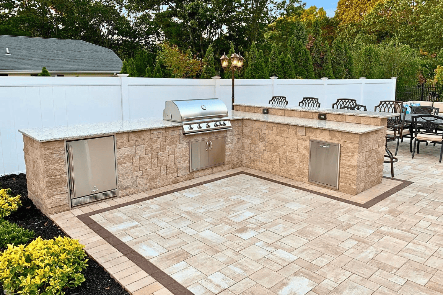 Outdoor Kitchens feature built-in stainless steel grills, storage cabinets, and stone countertops set on paver flooring. This inviting patio kitchen is complemented by chairs and lush greenery in the background.