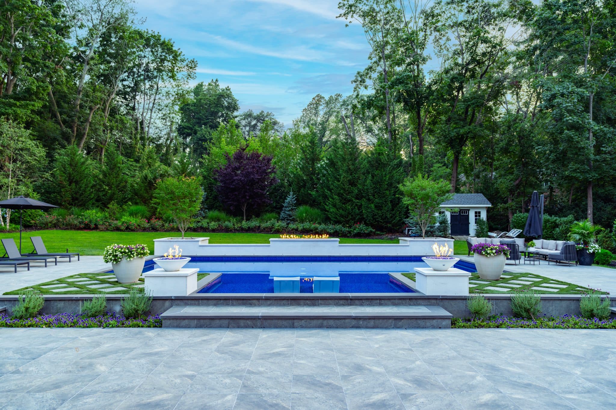 Modern backyard with a rectangular pool, lounge chairs, fire pit, manicured lawn, and surrounding trees under a blue sky.